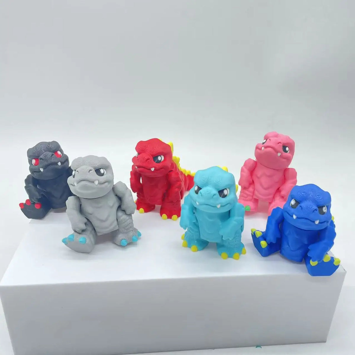 A collection of five 3D printed Godzilla sitting figures in various colors including black, gray, red, pink, and blue.