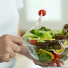 A hand holds a clear plastic dessert fork poised above a fresh salad with tomatoes and greens.