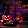 A glowing Halloween scene featuring a carved pumpkin, a birch tree with purple LED lights, and a skull on a wooden table.