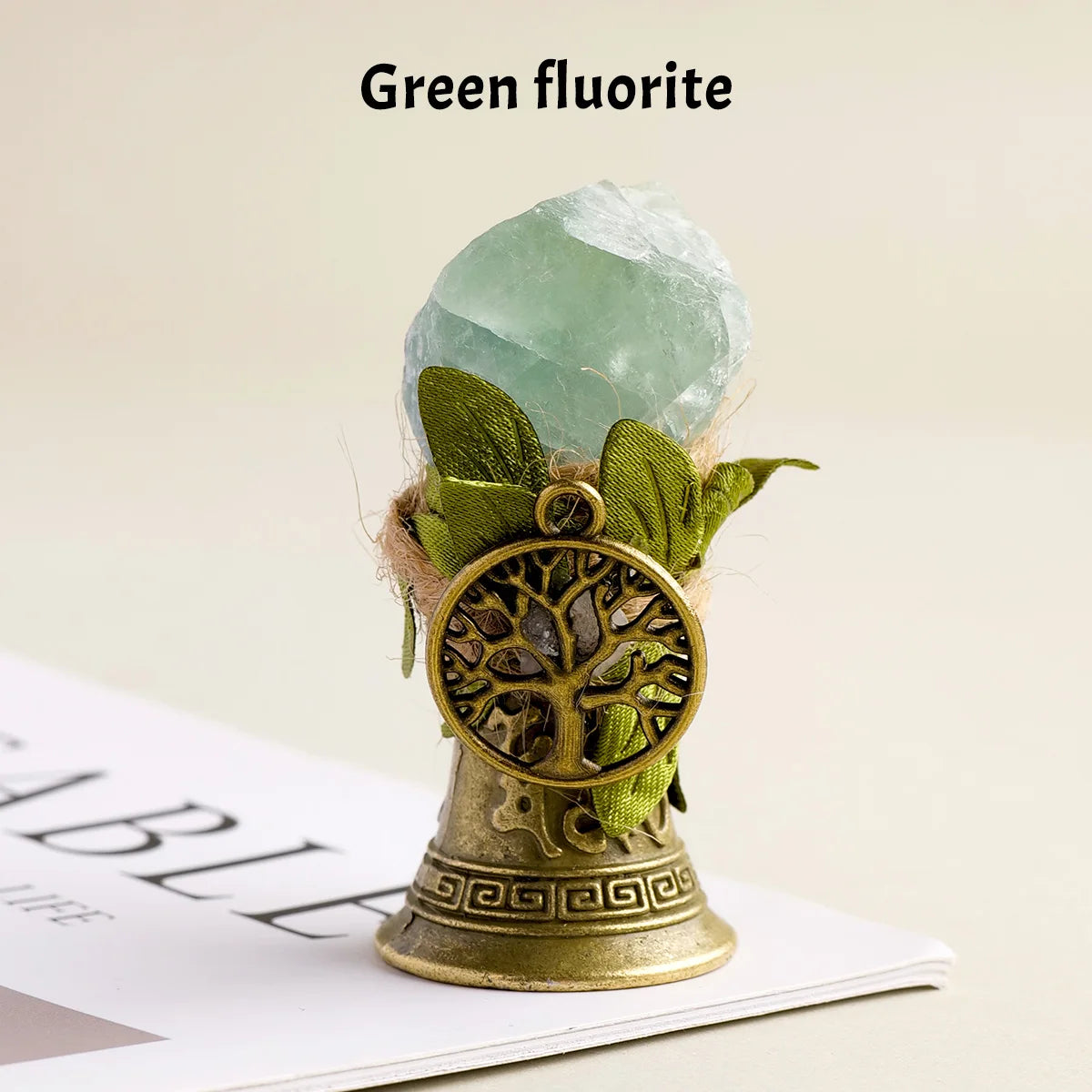 A green fluorite crystal is displayed in a brass bell with a copper finish and green foliage.