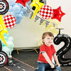 A toddler sits on a red racing car while a balloon arch decoration featuring black, yellow, and red stars and checkered patte