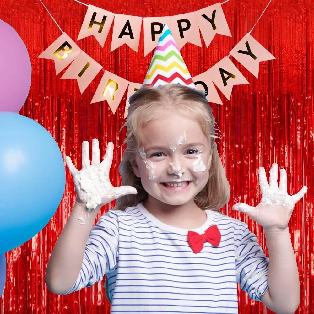 A smiling young girl with white frosting on her hands is in front of a red metallic foil fringe curtain birthday banner.