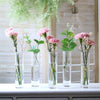 A set of twelve clear glass cylinder vases filled with greenery and delicate flowers arranged on a white surface.