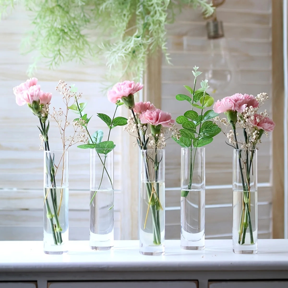 A set of twelve clear glass cylinder vases filled with greenery and delicate flowers arranged on a white surface.
