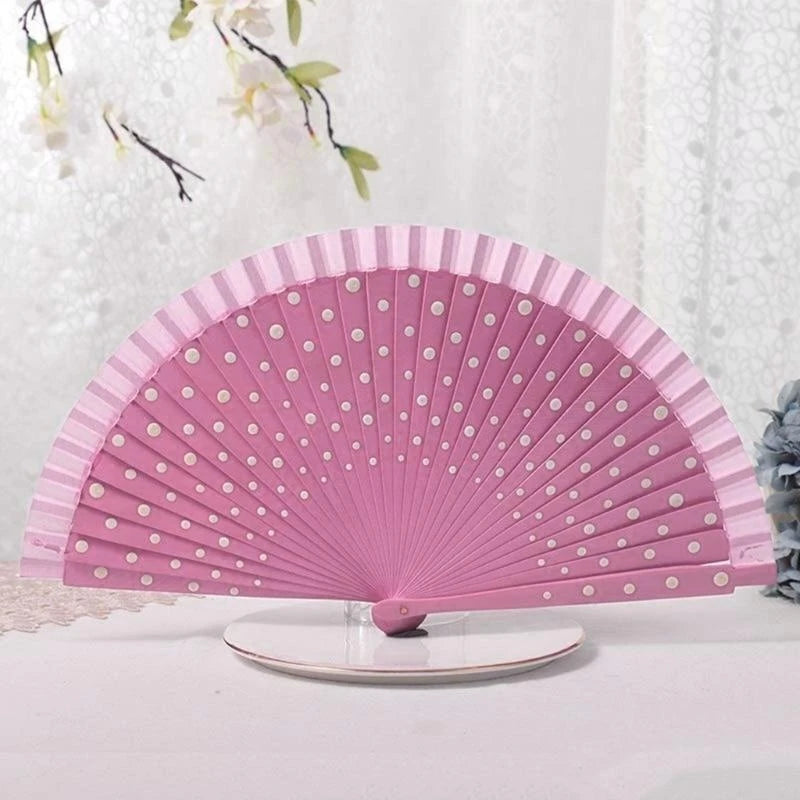 A pink wooden hand fan with white polka dots is displayed on a white surface.