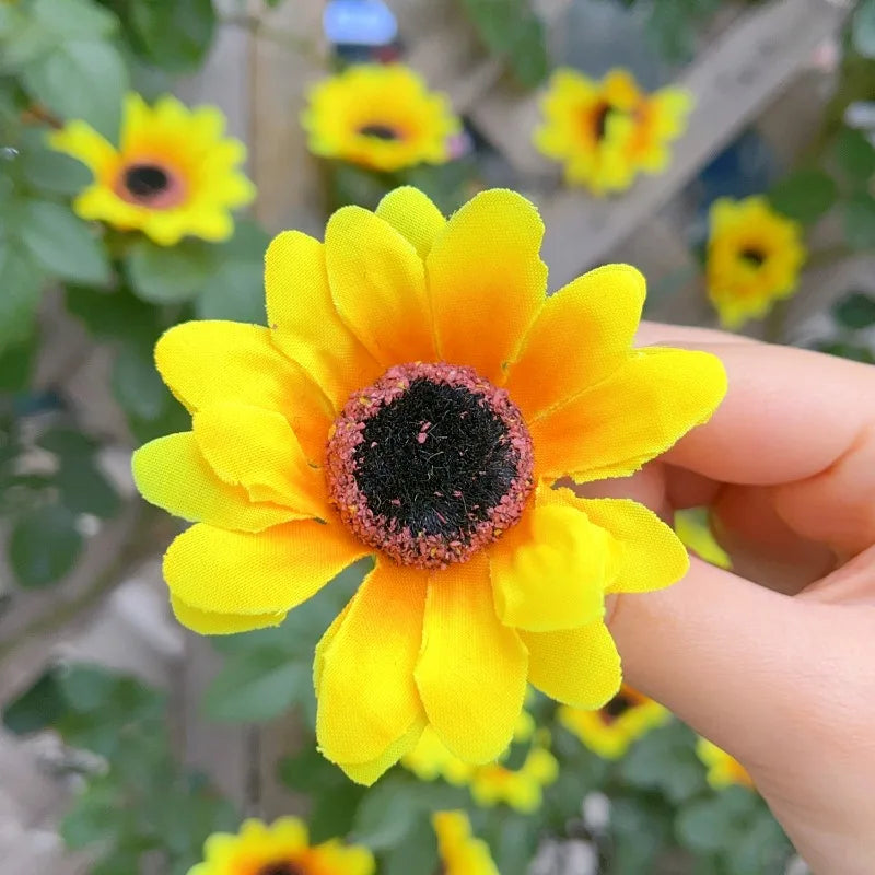 A hand holds a bright yellow artificial sunflower head with a dark center against a blurred green background.