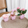 Three pink artificial roses in potted plants arranged on a woven mat.