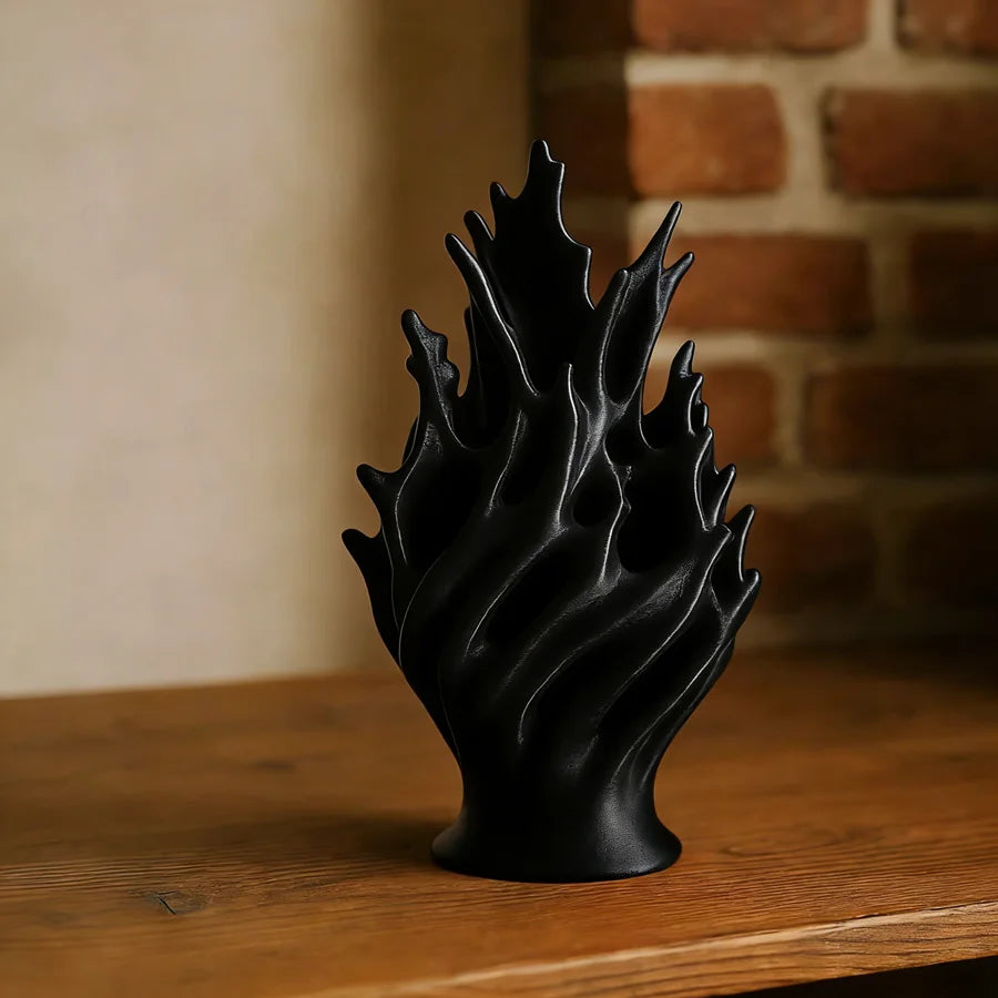 A black, sculptural plastic flower vase with ruffled leaves sits on a wooden surface next to a brick wall.