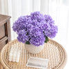 A bouquet of purple artificial hydrangea silk flowers in a light gray pot on a woven wooden tray.