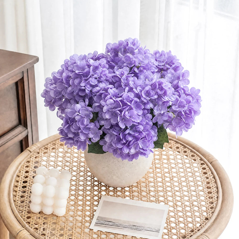 A bouquet of purple artificial hydrangea silk flowers in a light gray pot on a woven wooden tray.