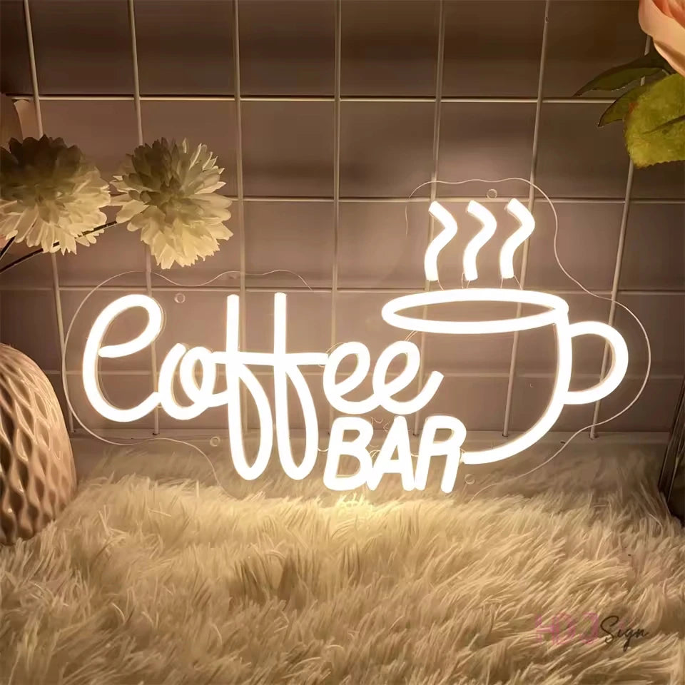 A white neon LED wall sign shaped like a coffee cup with steam, mounted on a tiled wall above a fluffy white surface.