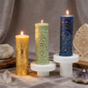 Three pillar candles in gold, green, and blue with celestial symbols displayed on a cloth.