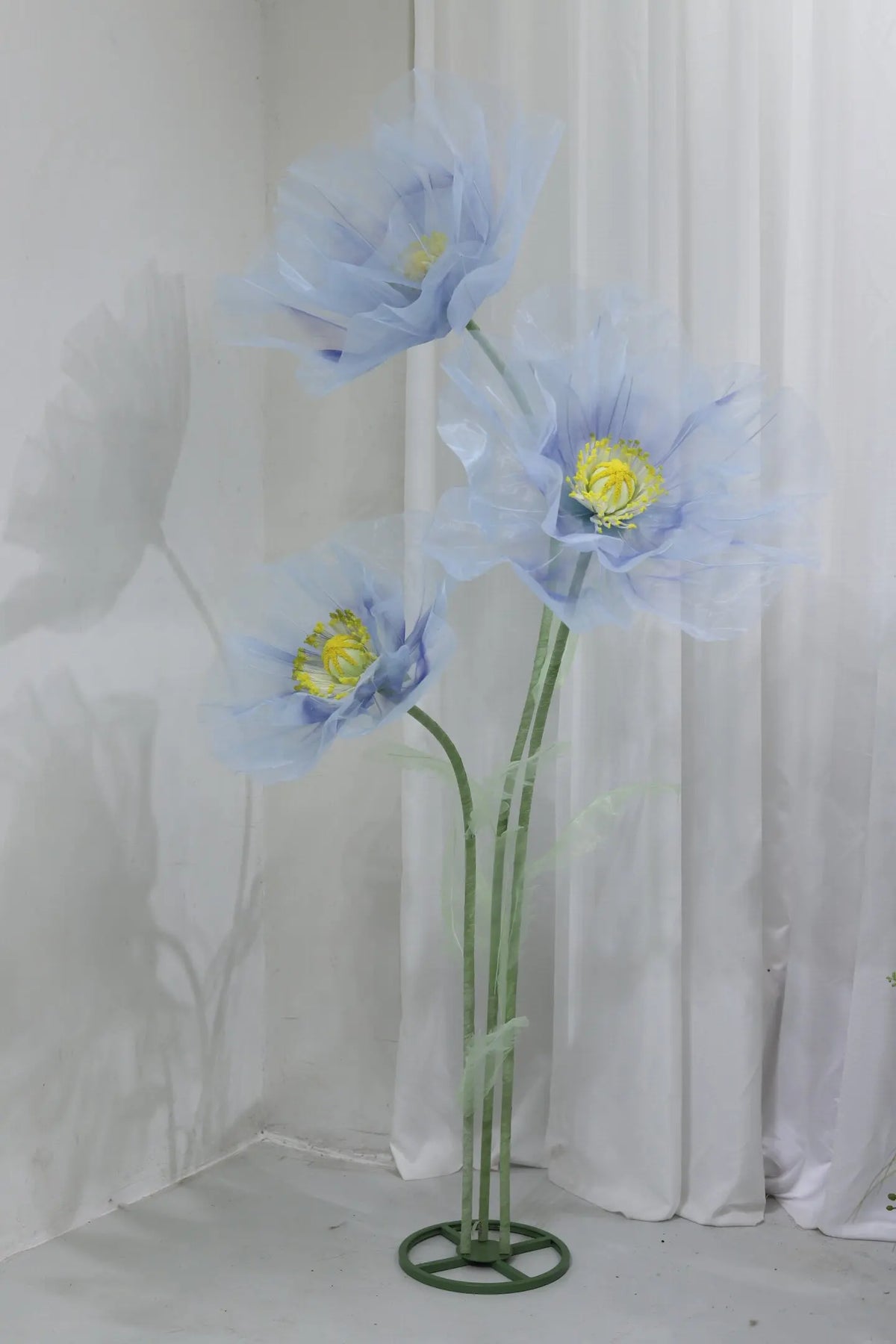 A large artificial silk blue poppy flower displayed on a green stand against a white curtain backdrop.