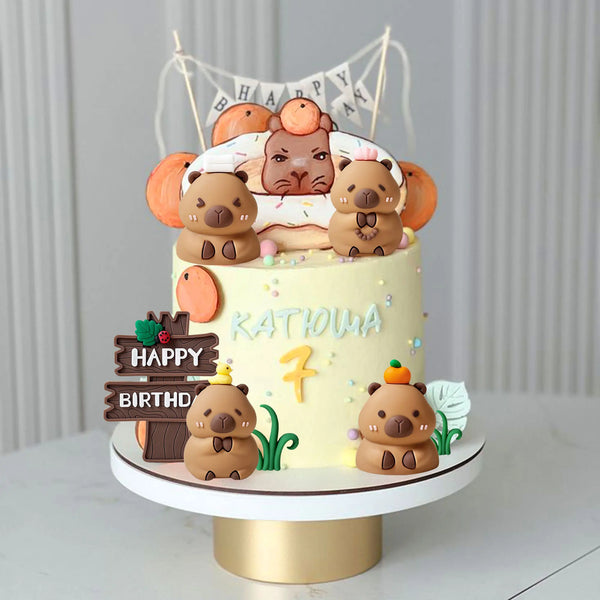 A tiered cake decorated with capybara animal figures and a banner, suitable for a birthday party.