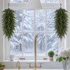 A golden teardrop swag of evergreen needles hangs in front of a bright window with snow outside.