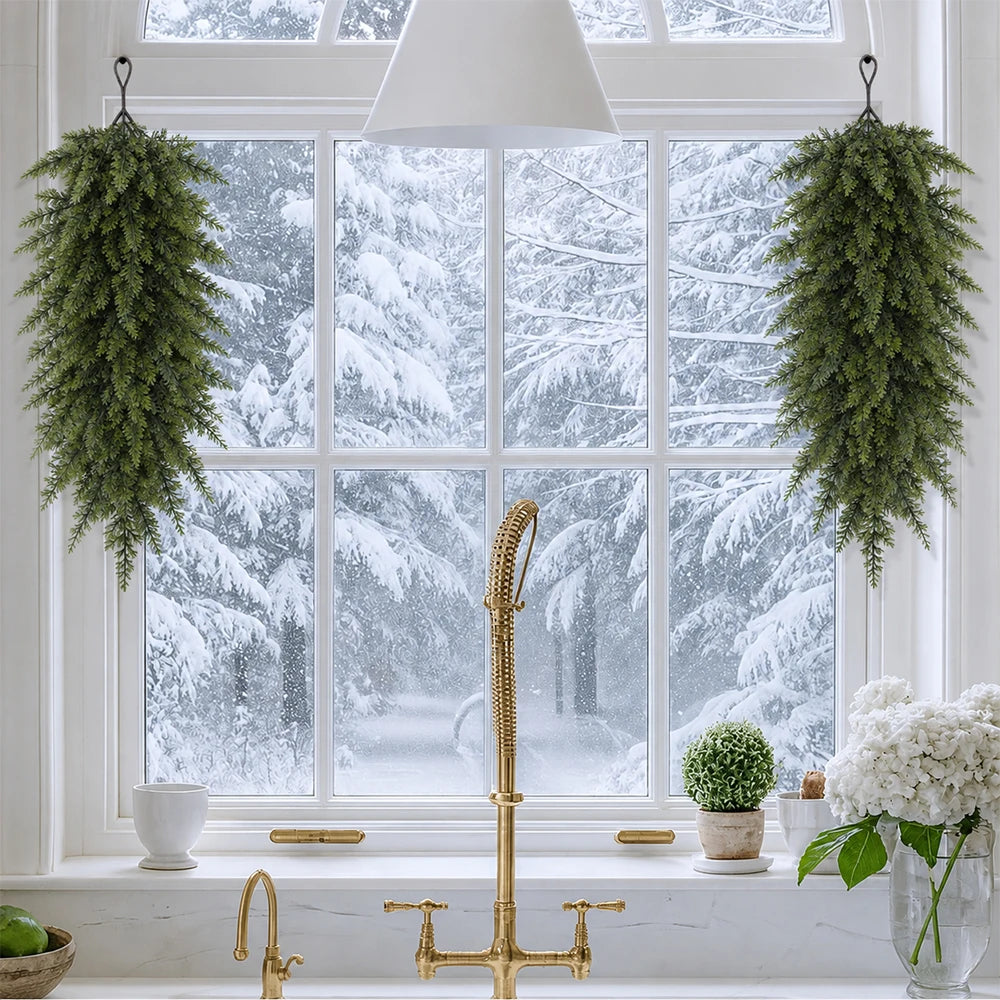 A golden teardrop swag of evergreen needles hangs in front of a bright window with snow outside.