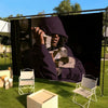 A person wearing a dark purple hood is seated in front of a large black backdrop outdoors.