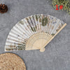 A hand carved bamboo folding fan with a natural, muted green and beige floral pattern is displayed on a gray surface.