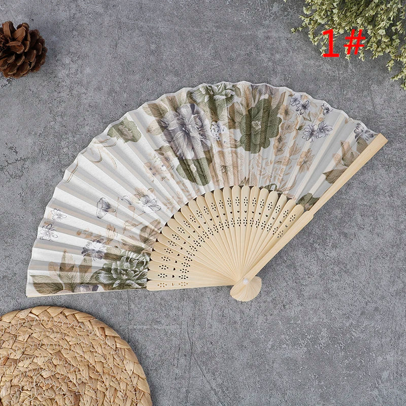 A hand carved bamboo folding fan with a natural, muted green and beige floral pattern is displayed on a gray surface.