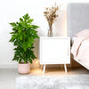 A potted artificial papaya leaf plant sits on a white nightstand next to a bed.