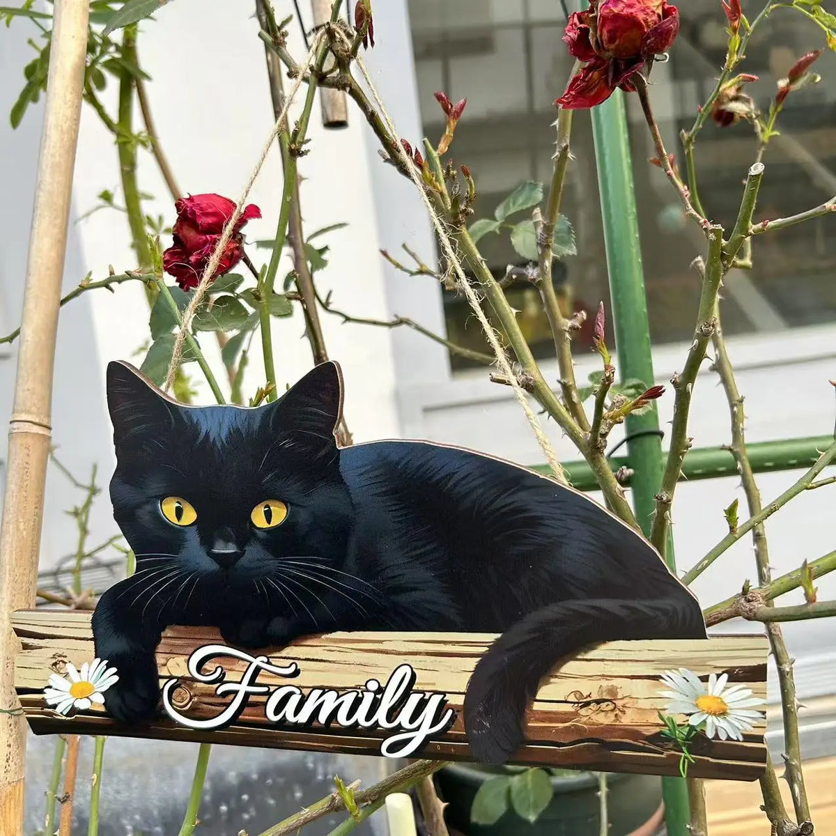 A black cat sits on a rustic wooden sign with the word Family carved into it, surrounded by green vines and red flowers.