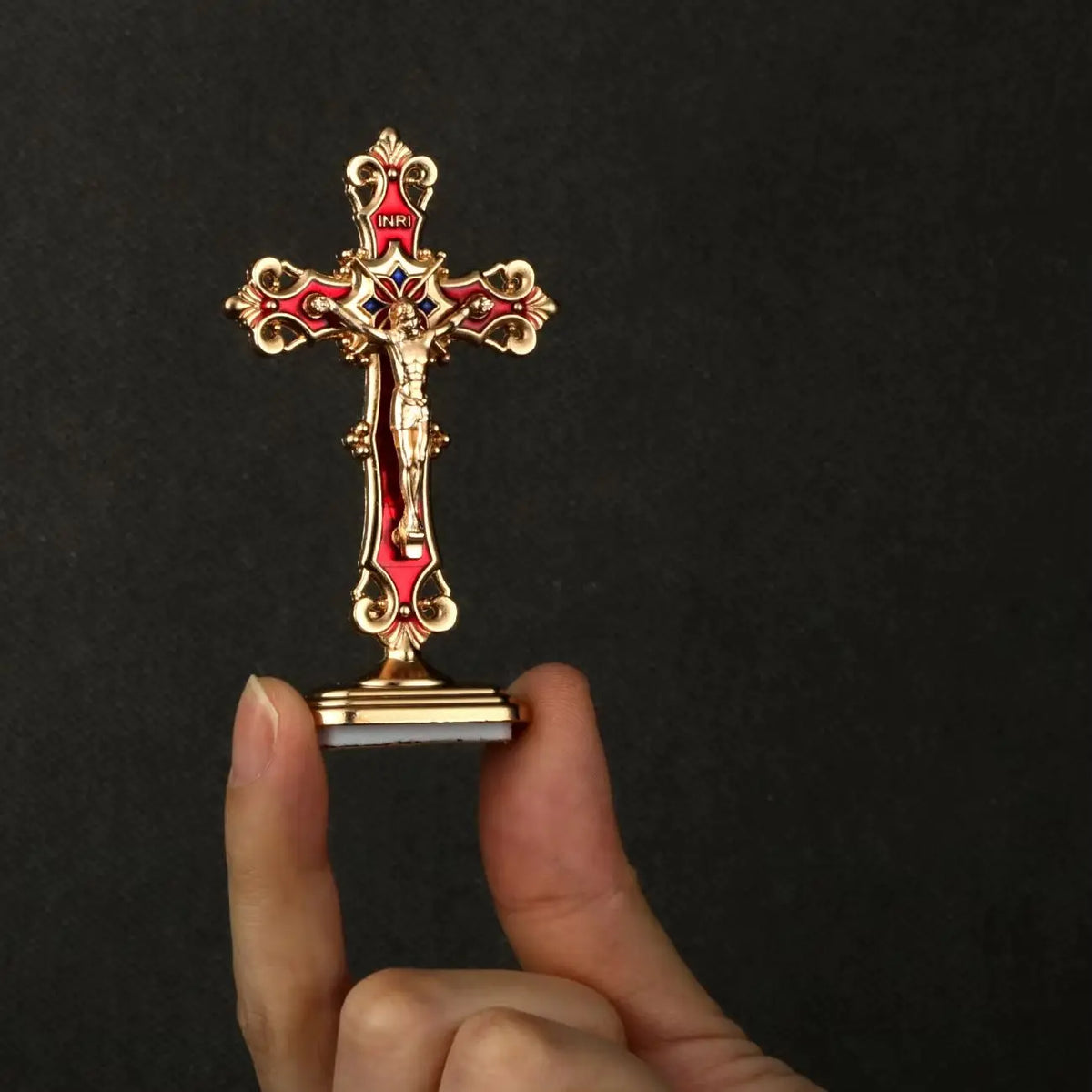 A gold metal crucifix cross with a red enamel figure and a white adhesive base being held by a hand.