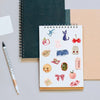 A spiral-bound notebook filled with watercolor dairy stickers, including animals and food items, next to a pencil.