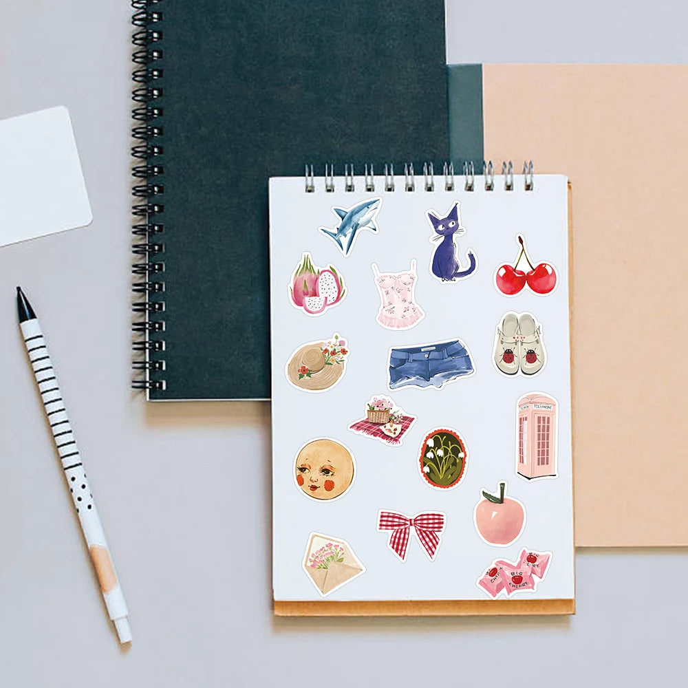 A spiral-bound notebook filled with watercolor dairy stickers, including animals and food items, next to a pencil.
