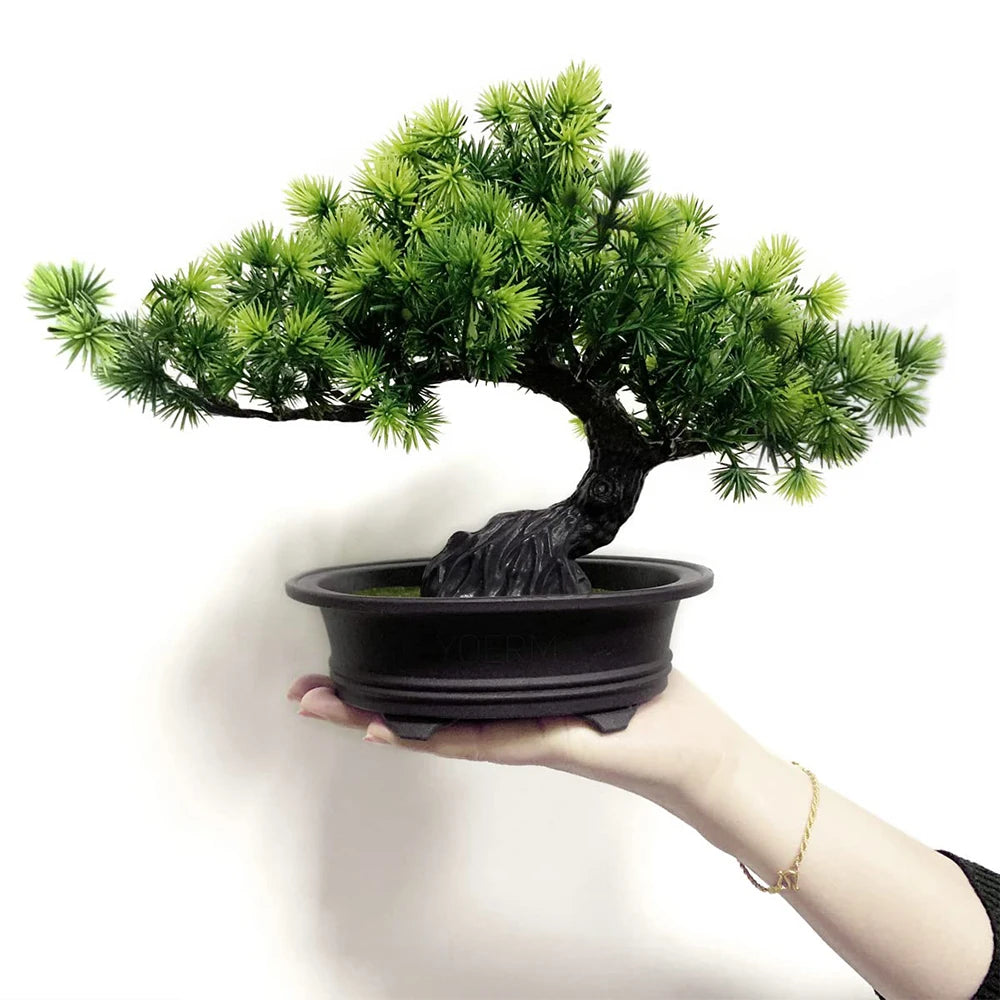 A hand holds an artificial bonsai tree with dense green foliage in a black pot against a white background.