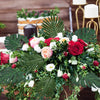 A lush floral arrangement featuring red and pink roses, white flowers, and large green artificial tropical palm leaves for pa