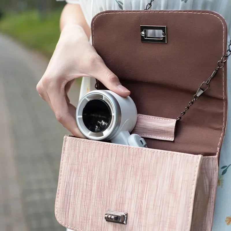 A hand is holding a foldable mini handheld fan with a USB charging cable inside a brown and pink fabric pouch.