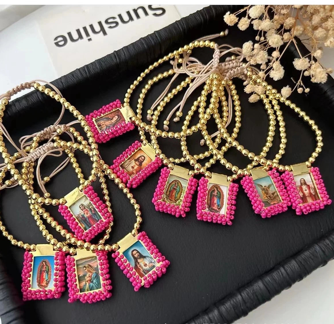 Gold-plated charm bracelets featuring Virgin Mary Guadalupe figures and pink beadwork displayed on a black tray.