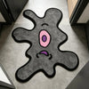 A gray cartoon monster rug with pink eyes and purple mouth is displayed on a light-colored floor.