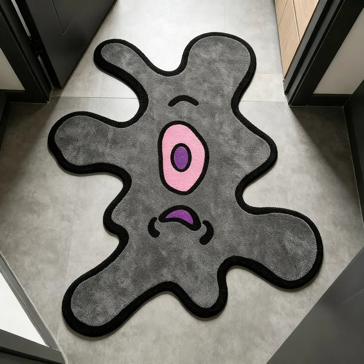 A gray cartoon monster rug with pink eyes and purple mouth is displayed on a light-colored floor.