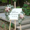 A wooden sign with 'Welcome Grace & Daniel 10.20.24' and floral arrangements featuring dusty rose and blue eucalyptus.