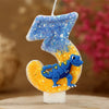 A birthday candle shaped like the number 3, decorated with blue glitter and a yellow dinosaur figure.