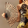 A woman holds a leopard print folding hand fan with a black frame against a neutral background.