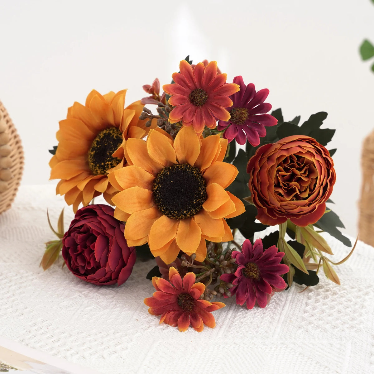 Un bouquet de tournesols artificiels et de pivoines rouges et oranges sur un tissu blanc.