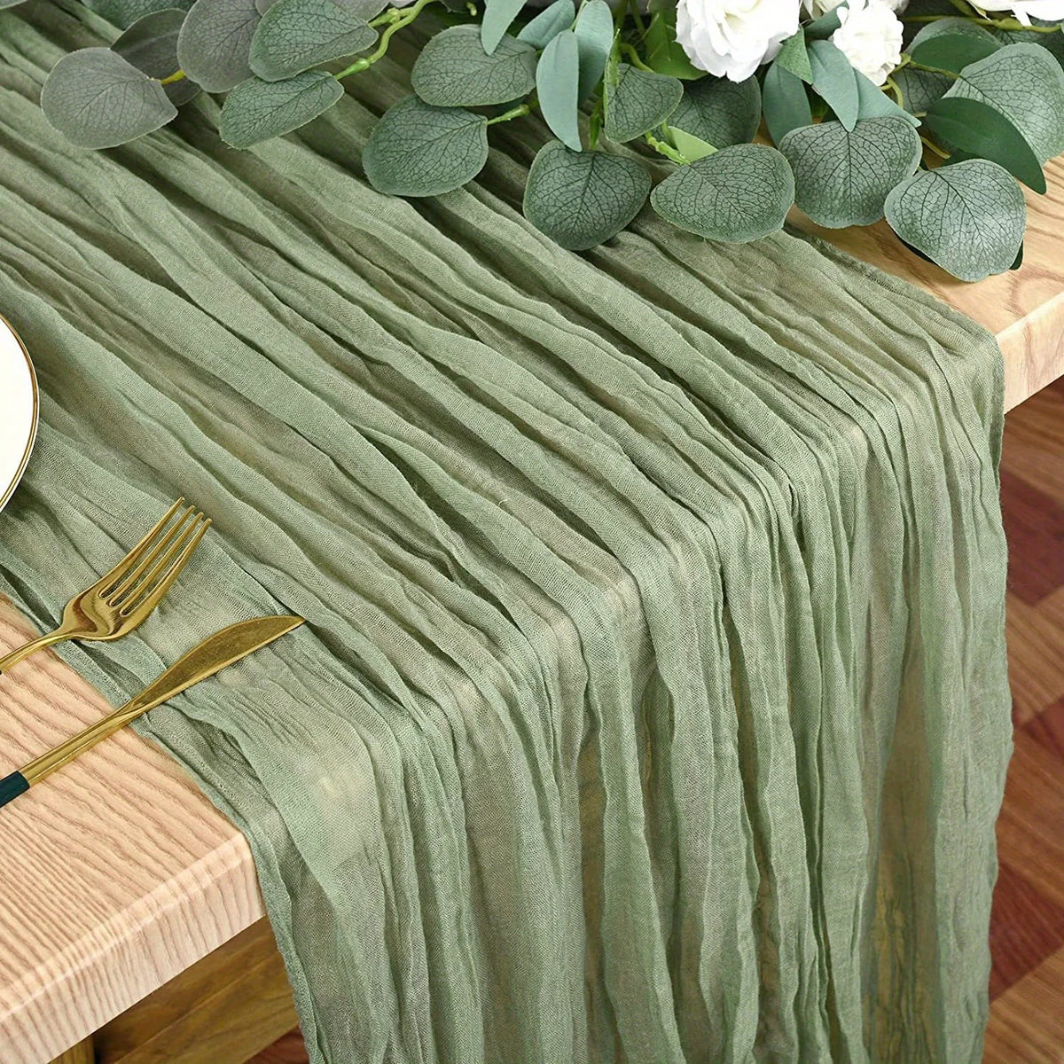 A sage green cheesecloth table runner draped over a wooden table with gold cutlery and white flowers.