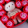 A pink teddy bear is nestled among red and pink rose flowers in a gift box.