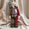 A standing figurine of a bear with a pink face, brown body, and red clothing, shown against a white fabric background.