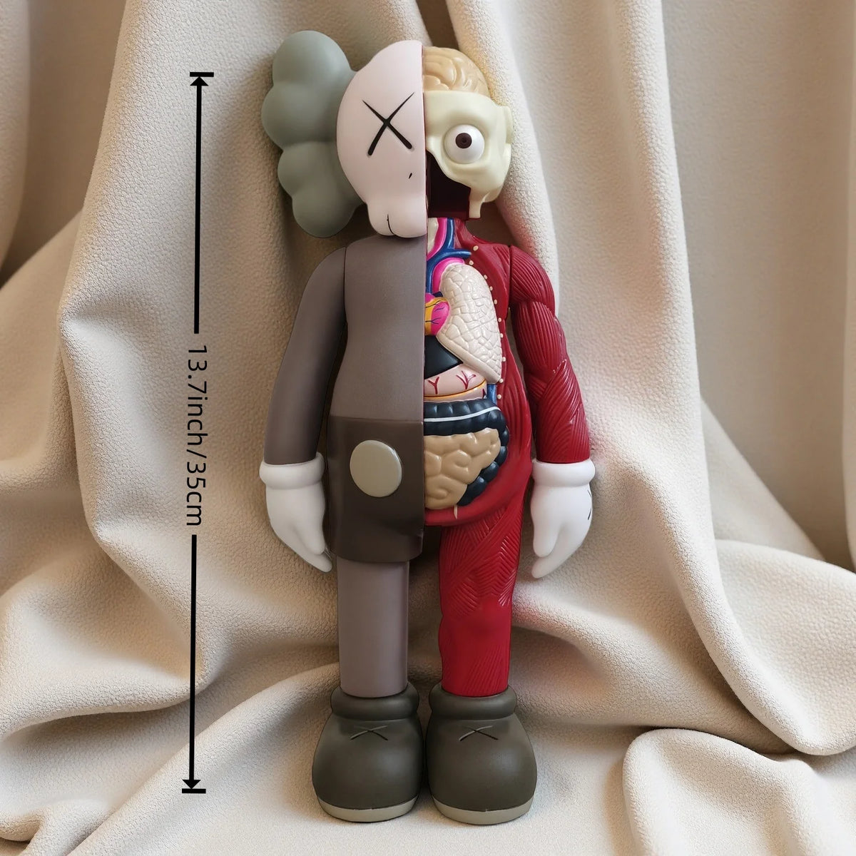 A standing figurine of a bear with a pink face, brown body, and red clothing, shown against a white fabric background.