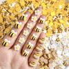 A hand displays a set of cute bee and daisy 3D resin nail art charms in various colors on a yellow background.