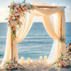 Arche de mariage décorée de draperies en mousseline crème et de compositions florales sur fond de mer bleue.