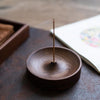 A dark brown solid wood incense stick burner with a thin metal stick resting on a dark slate surface.