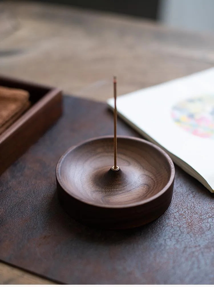 A dark brown solid wood incense stick burner with a thin metal stick resting on a dark slate surface.