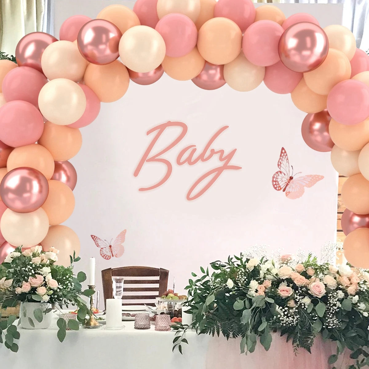 A pink and peach balloon arch decoration for a baby shower or party with floral greenery and pink accents.