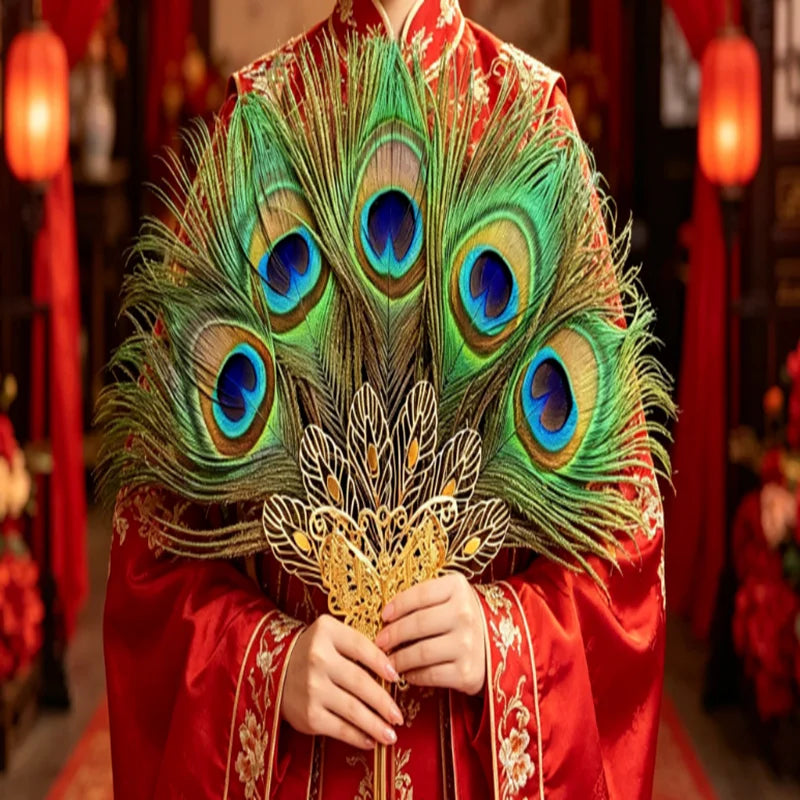 Une personne tient un éventail de plumes de paon naturel avec des plumes vertes et bleues irisées contre un vêtement rouge.