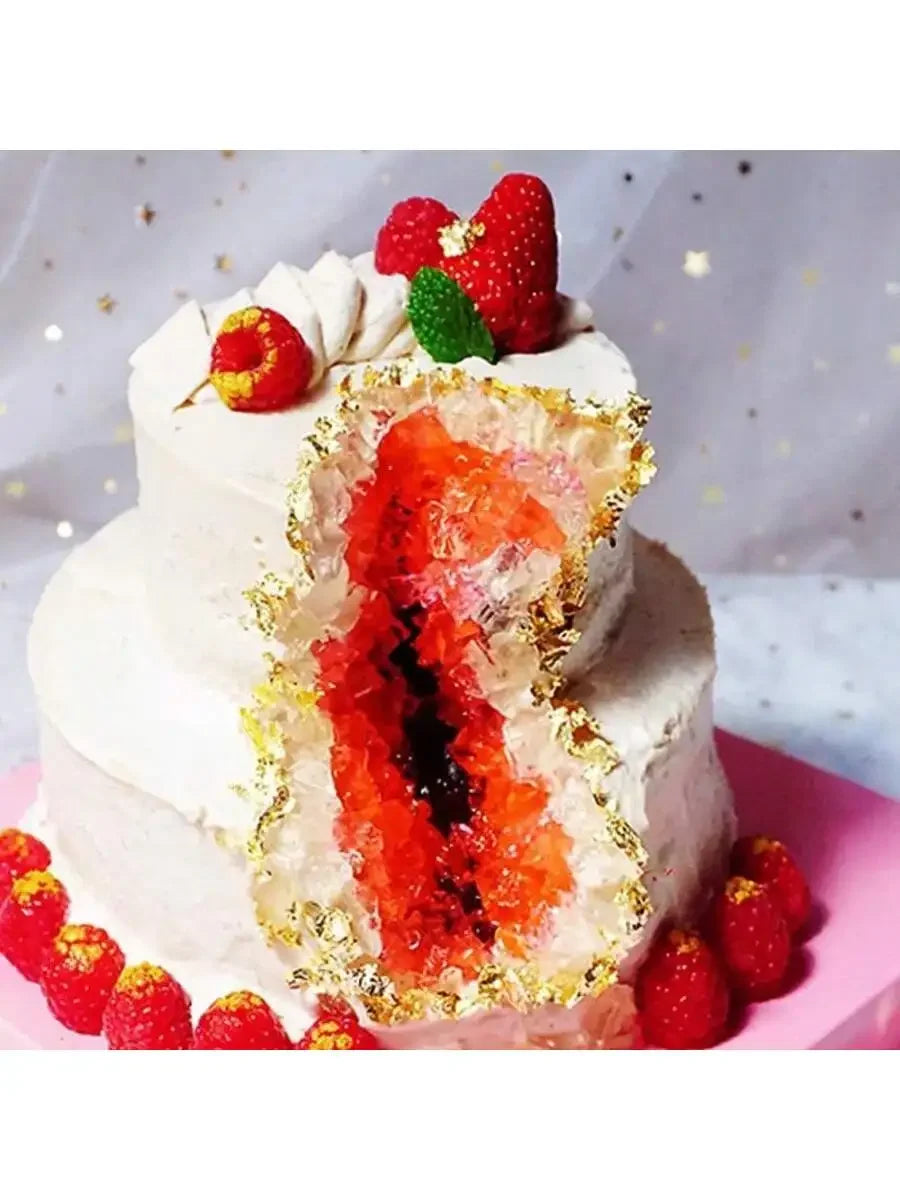 A white layer cake with a slice removed, revealing bright red fruit and gold leaf flakes for decoration.