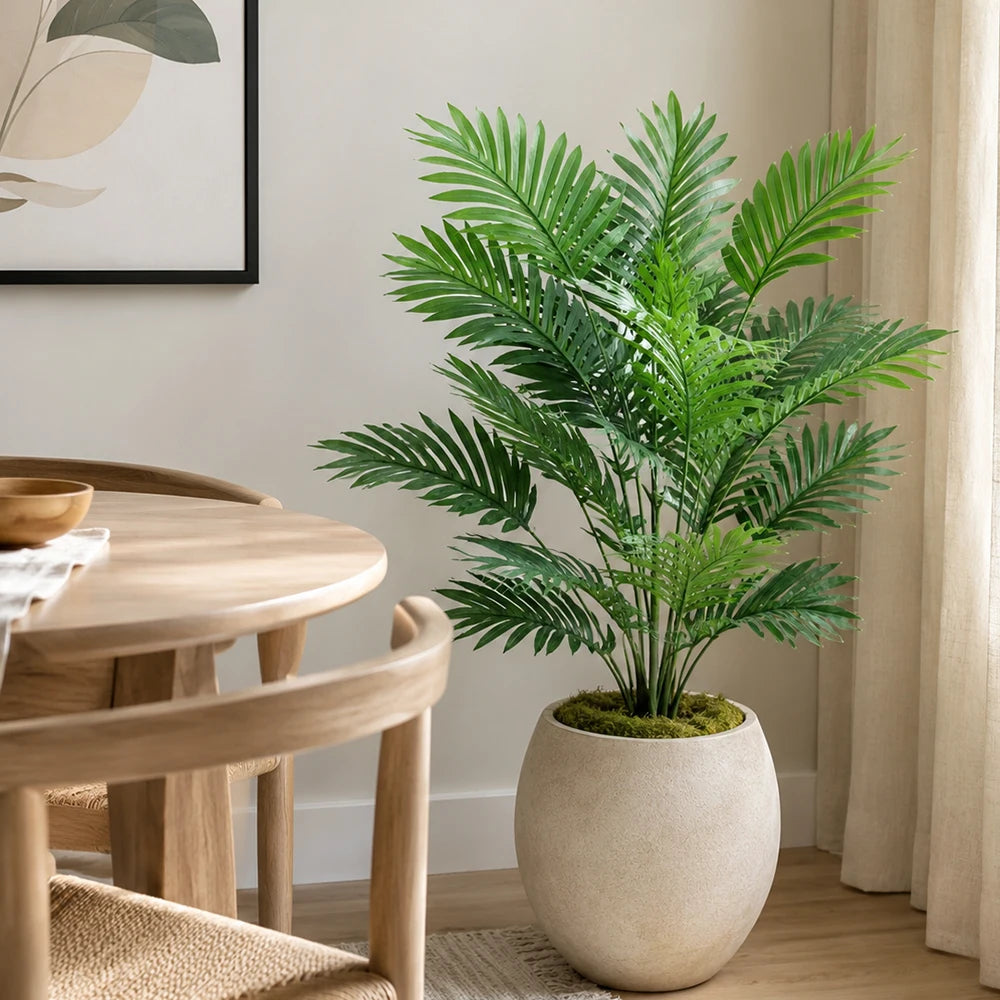 A vibrant artificial areca palm tree in a beige pot sits on a wooden table next to a window.